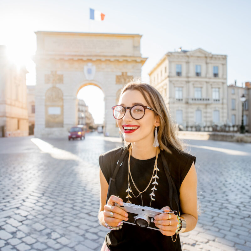 Portrait of a young happy woman tourist with photo camera in front of the famous Triumphal Arch during the morning light in Montpellier city, France